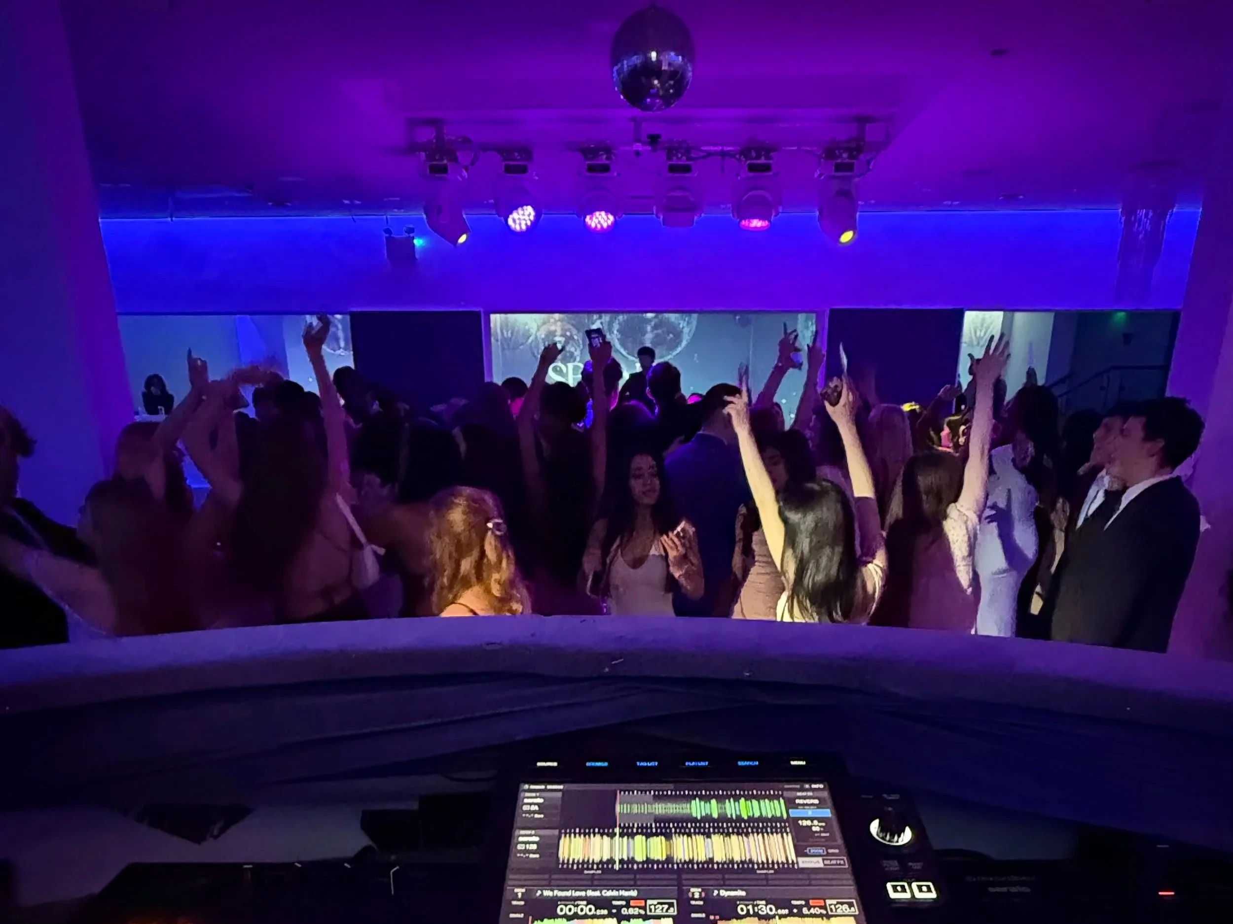 A DJ booth at a nightclub with a DJ console displaying colorful sound wave patterns, and a crowd of people dancing under purple and yellow stage lighting, with a disco ball hanging from the ceiling.