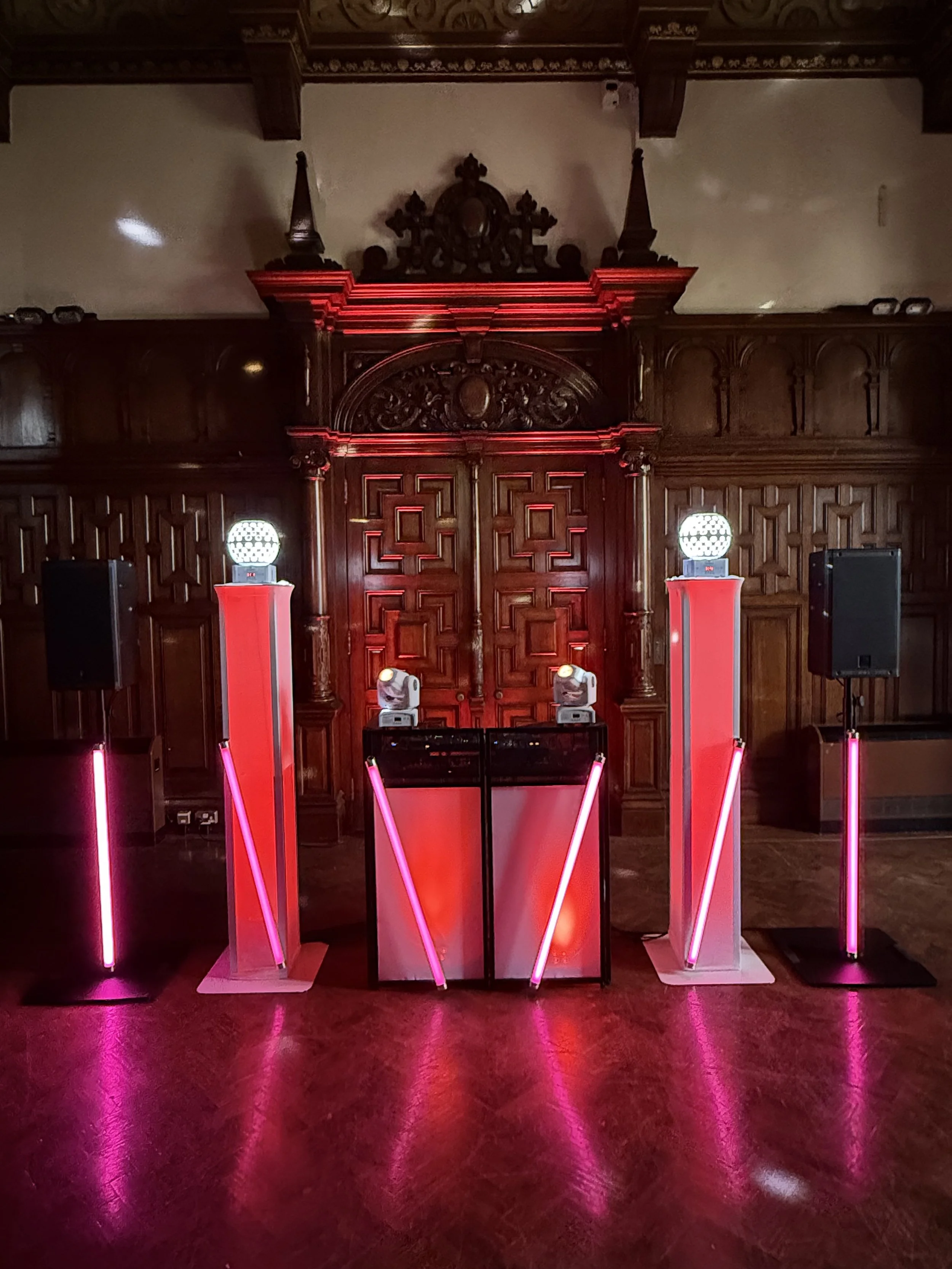 An indoor event space with dark wooden wall paneling and ornate woodwork on the ceiling. Three tall pink illuminated light fixtures are arranged in a row, with additional pink and white lighting effects on the floor. Two black speakers are positioned on either side of the setup.
