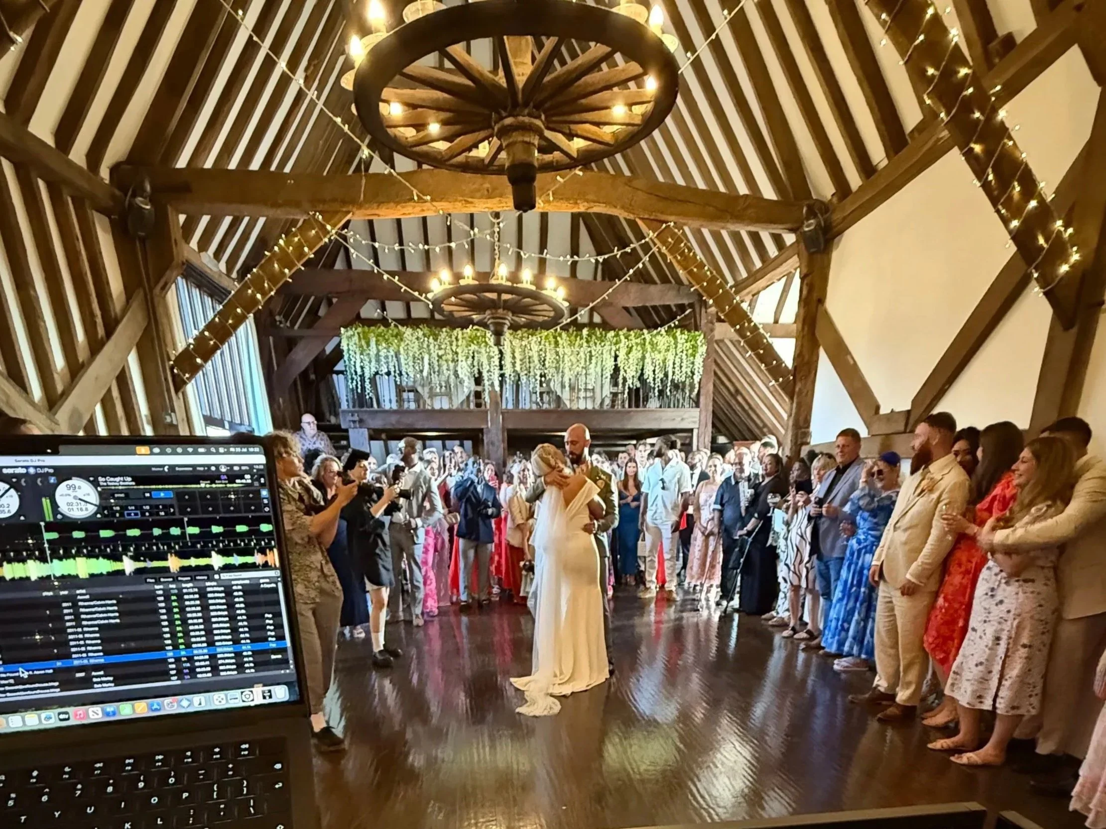 Indoor wedding reception with a bride and groom dancing in the center. Guests are standing around them, watching and smiling. The ceiling has wooden beams with hanging string lights and large wagon wheel chandeliers. A DJ or music setup is visible in the foreground.