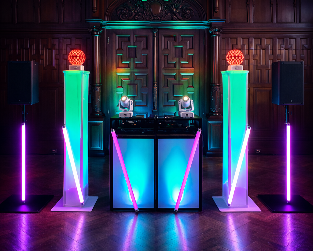 Colorful DJ setup with neon lights, two tall illuminated columns with speakers, a central DJ booth with lighting, and a dark wooden ornate wall background.