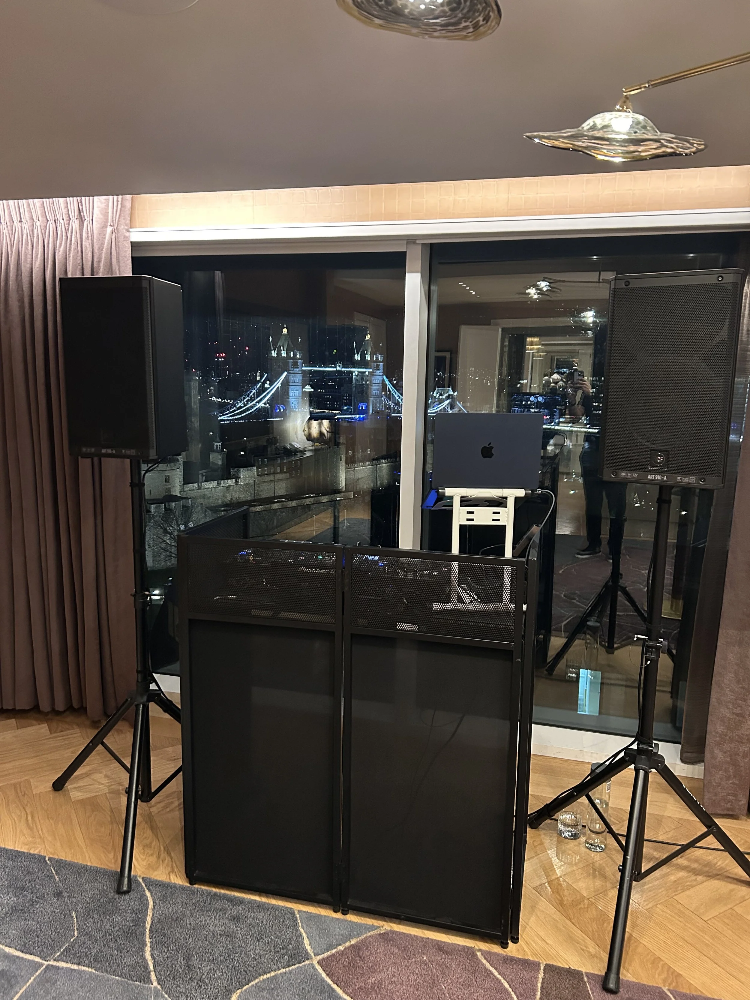 Indoor party or event setup with two large speakers on tripod stands, a small table with a computer, and a glass barrier in front. The background shows a cityscape through a large window, with bridge lights and tall buildings illuminated at night. The room has curtains on the left, ceiling lights, and a multicolored patterned rug on the wooden floor.