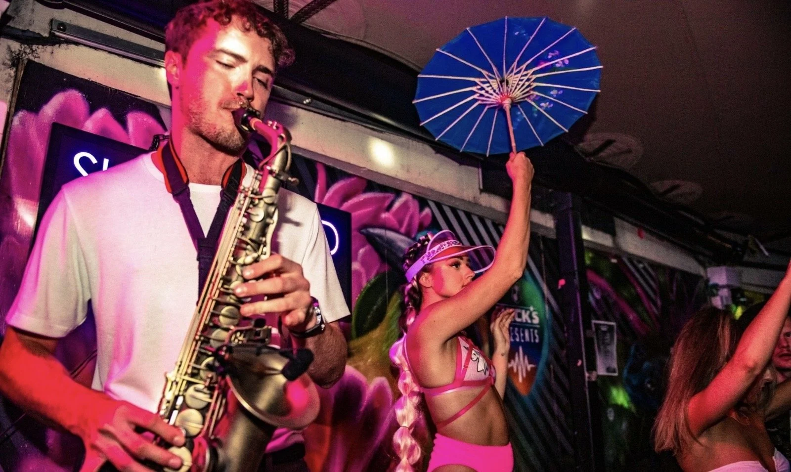 A man playing a saxophone with a woman holding a blue umbrella above her head, both dancing in a brightly lit colorful club or party setting.