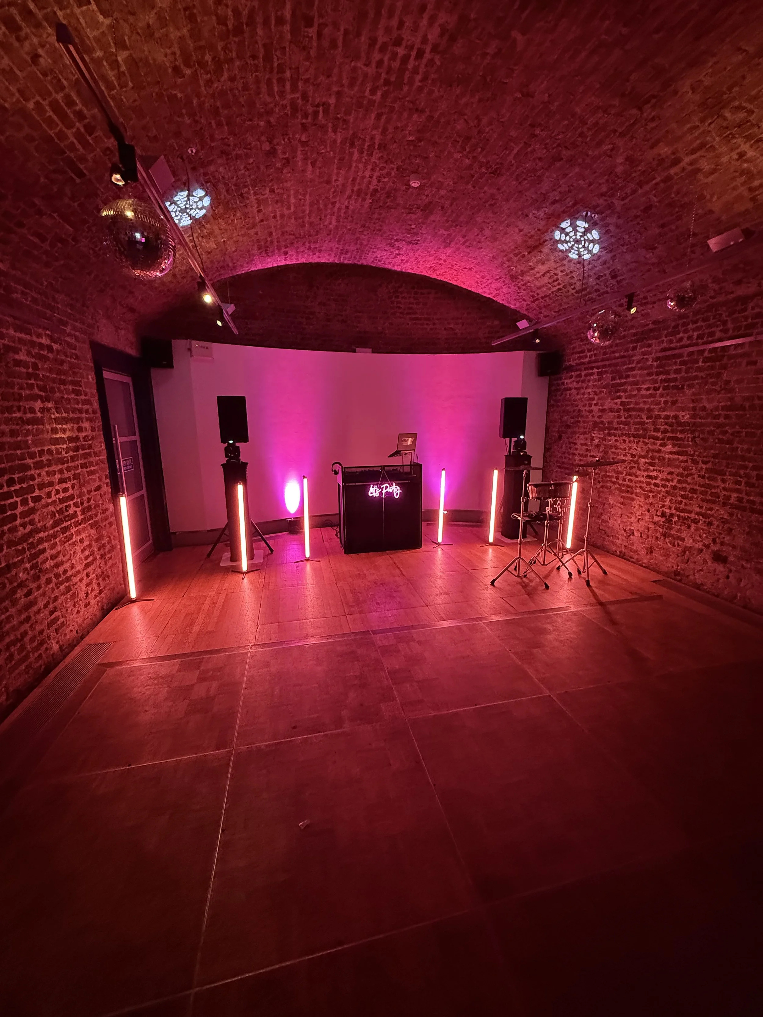 An indoor event space with brick walls and a vaulted ceiling, decorated with pink lighting and disco balls, set up for a party or performance with speakers and lighting equipment.
