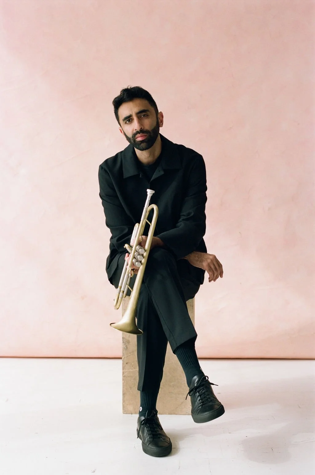 Riley sits on the edge of a platform wearing a dark suit. He holds a trumpet in his hands and leans toward the camera.