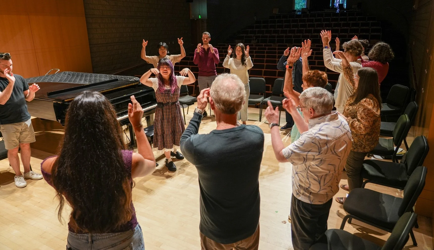 A music instructor leads a group of adult musicians. They are standing in a circle onstage by a grand piano.