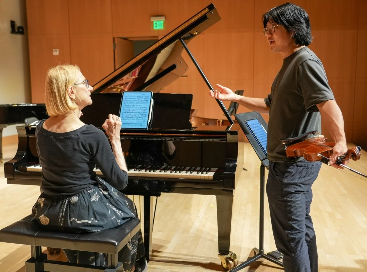 A woman sits at a grand piano on a stage, in conversation with a violinist standing next to her.