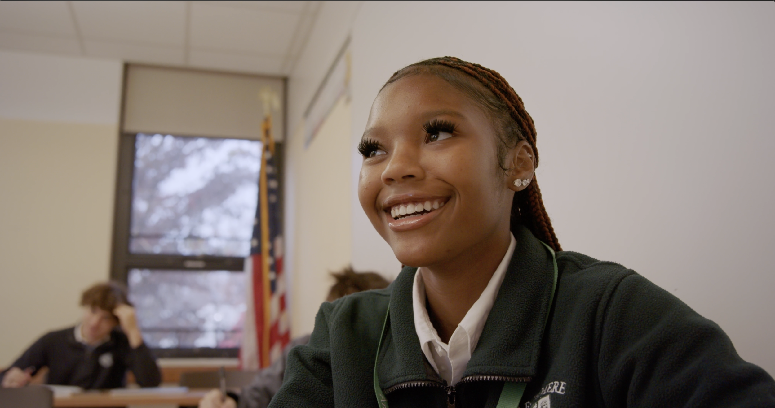 Young woman smiling in a classroom with students in the background, some looking down and others with head in hand, American flag and a window showing trees outside.