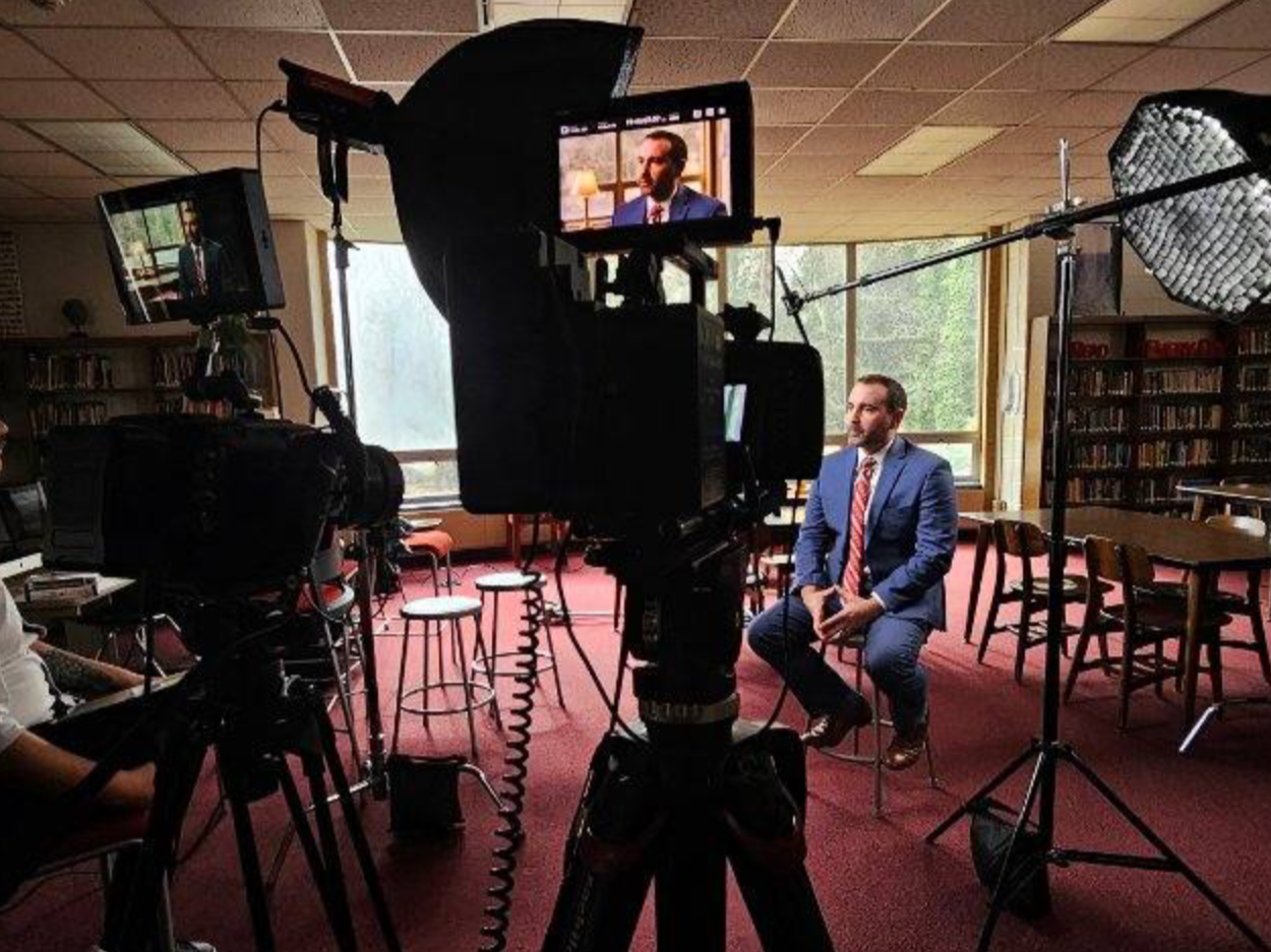 A man in a blue suit and red tie sitting on a stool is being filmed with multiple cameras and lighting equipment in a room with bookshelves and large windows.