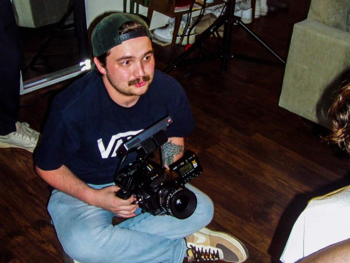 A man sitting on the floor holding a camera in a room with photography equipment, a large softbox light, a mirror, a plant, a cabinet, and a couch.