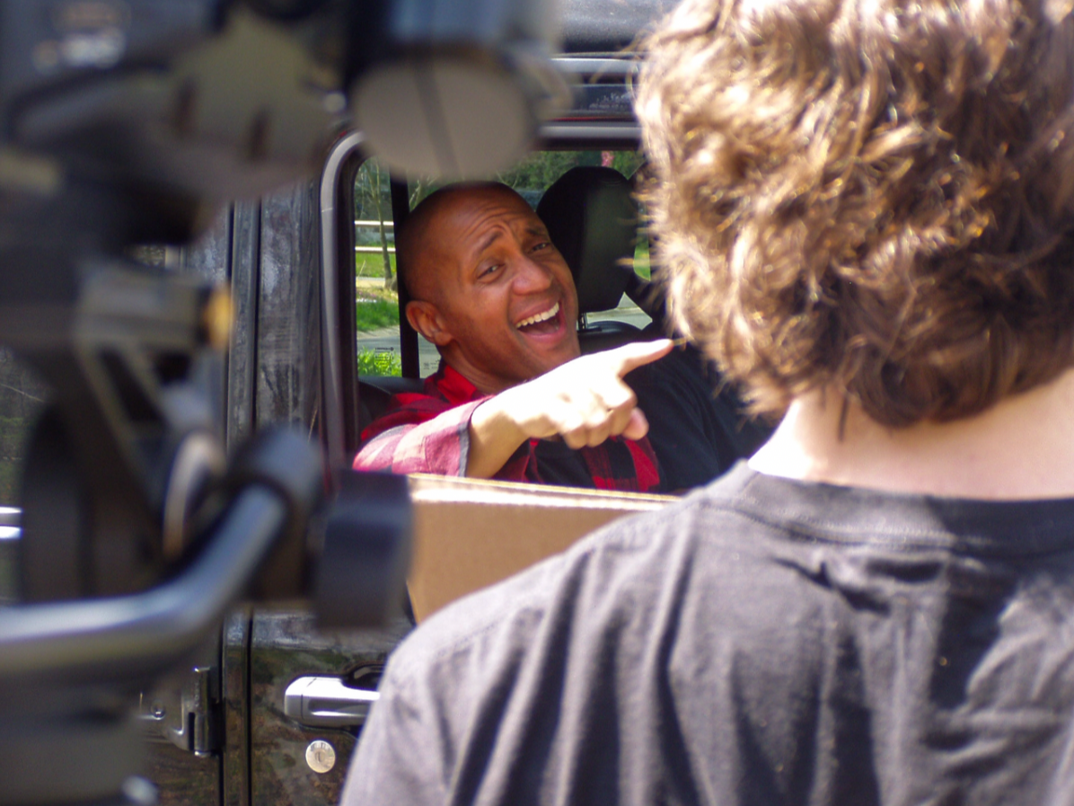 A man sitting in a vehicle pointing and smiling, as a person outside the vehicle with curly hair is seen from behind.