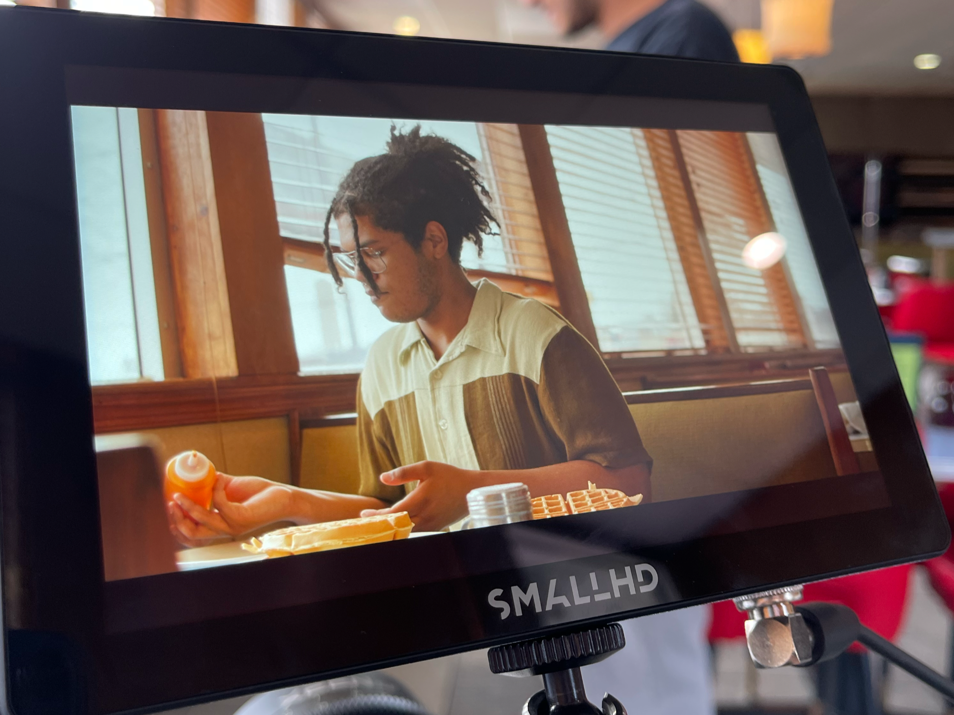 A camera monitor showing a young man with glasses and dreadlocks in a restaurant, holding a bottle of syrup next to waffles, with a man in a blue shirt in the background.