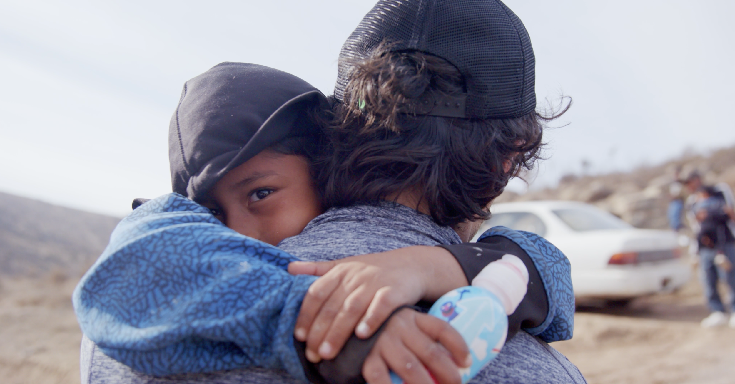 A child hugging an adult outdoors near a desert area, with a white car and a person on a motorcycle in the background.