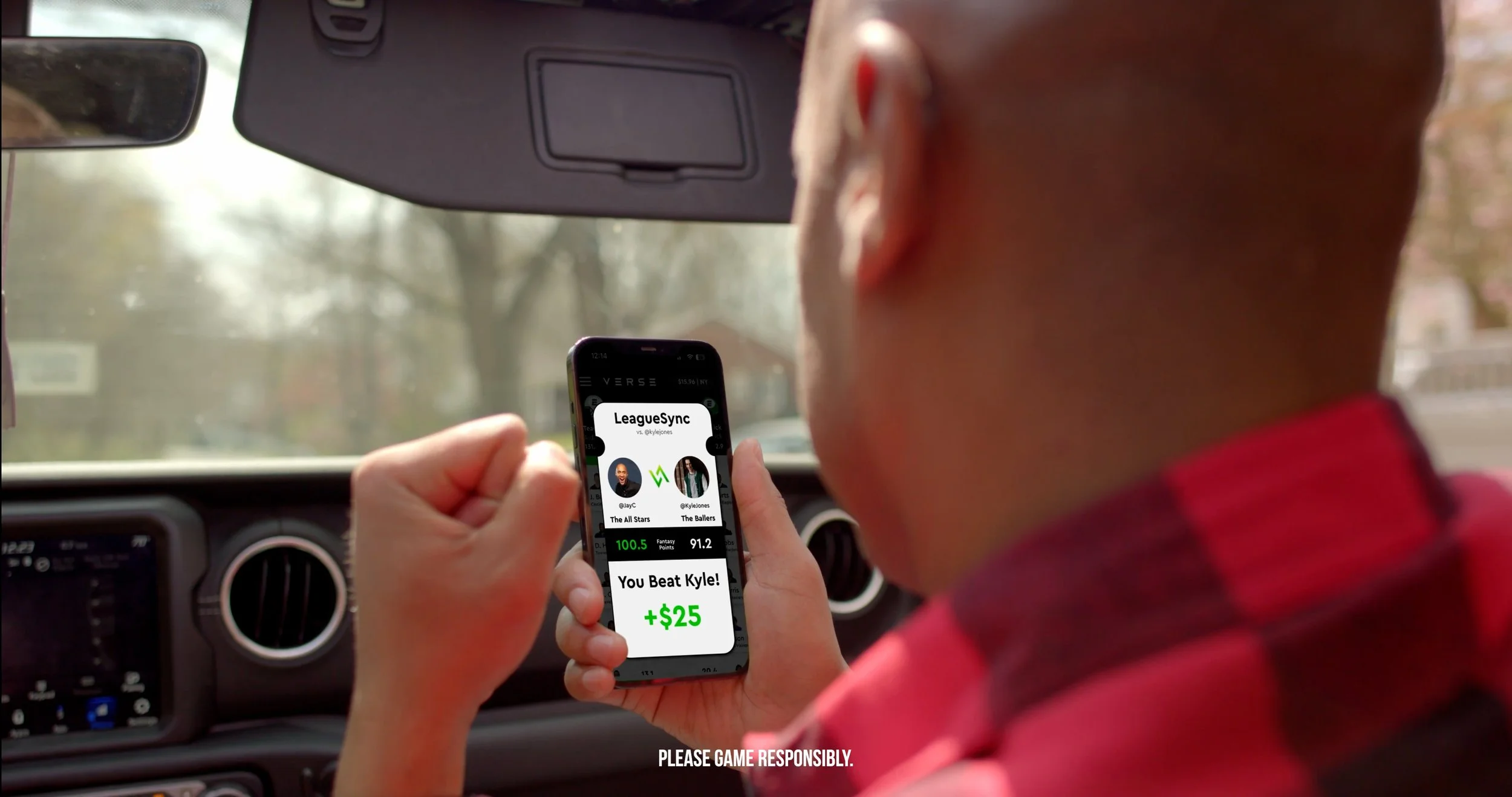 A man in a red plaid shirt sitting inside a car, holding a smartphone displaying a fantasy sports app showing he beat a player named Kyle and winning $25, with a blurred outdoor background visible through the windshield.