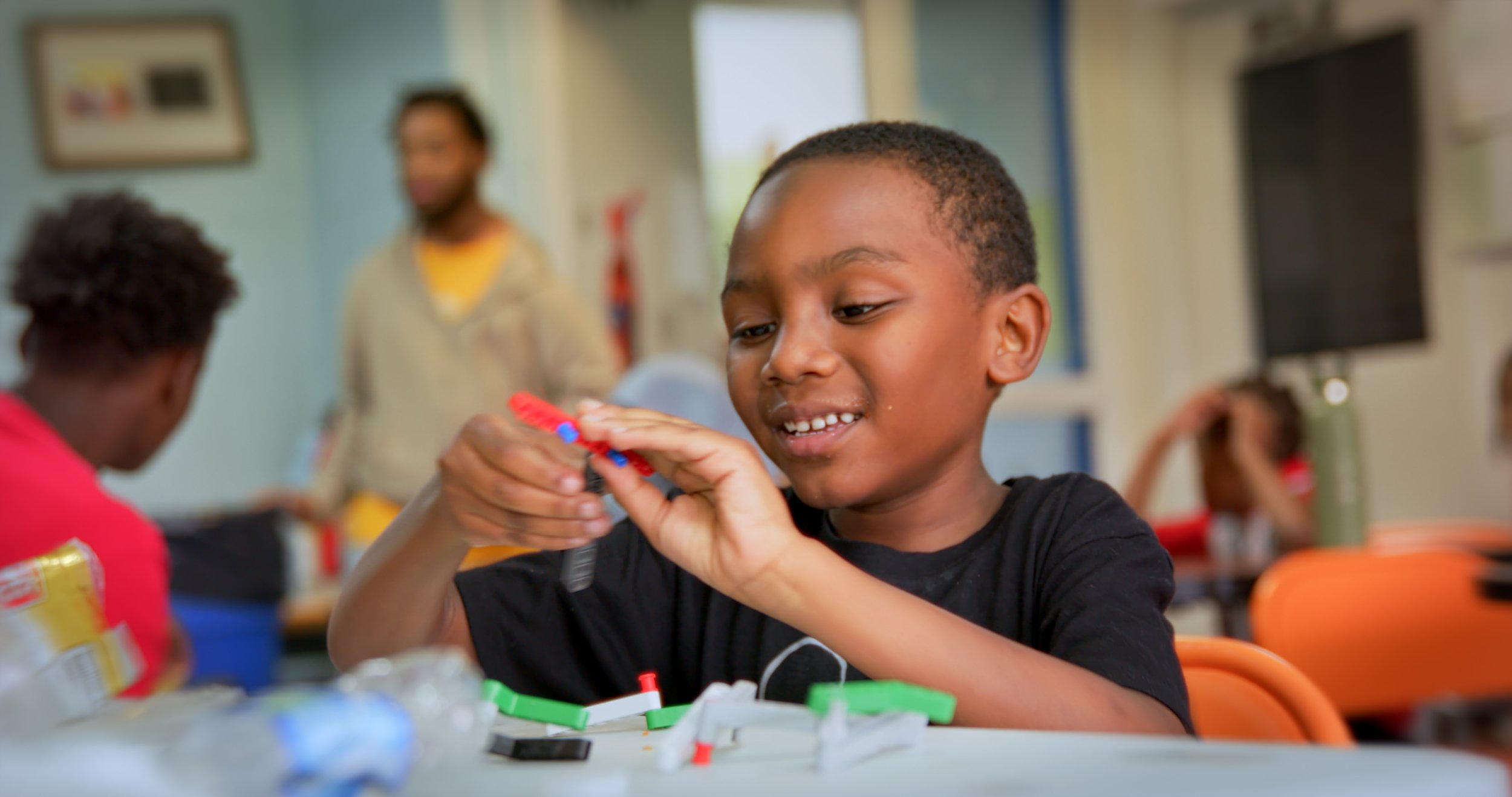 A young boy playing with building toys at a table in a classroom, with children and an adult in the background.