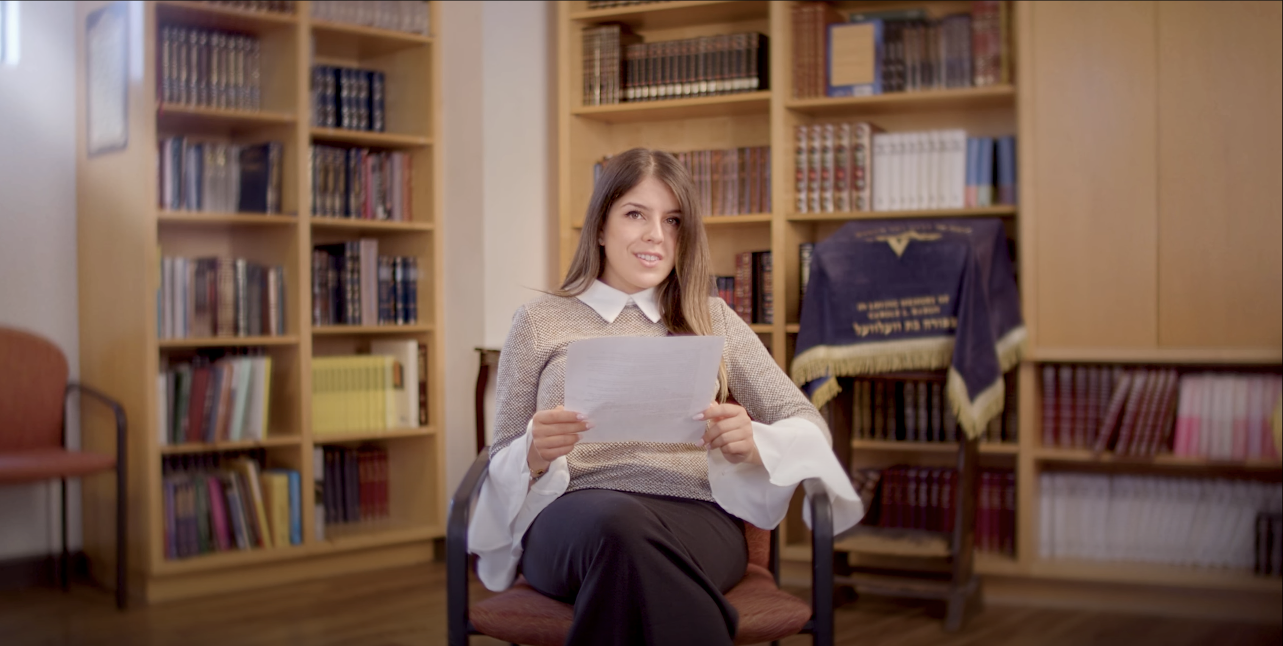 A woman with long brown hair, sitting in an office chair, holding a paper, smiling, in front of bookshelves filled with books.
