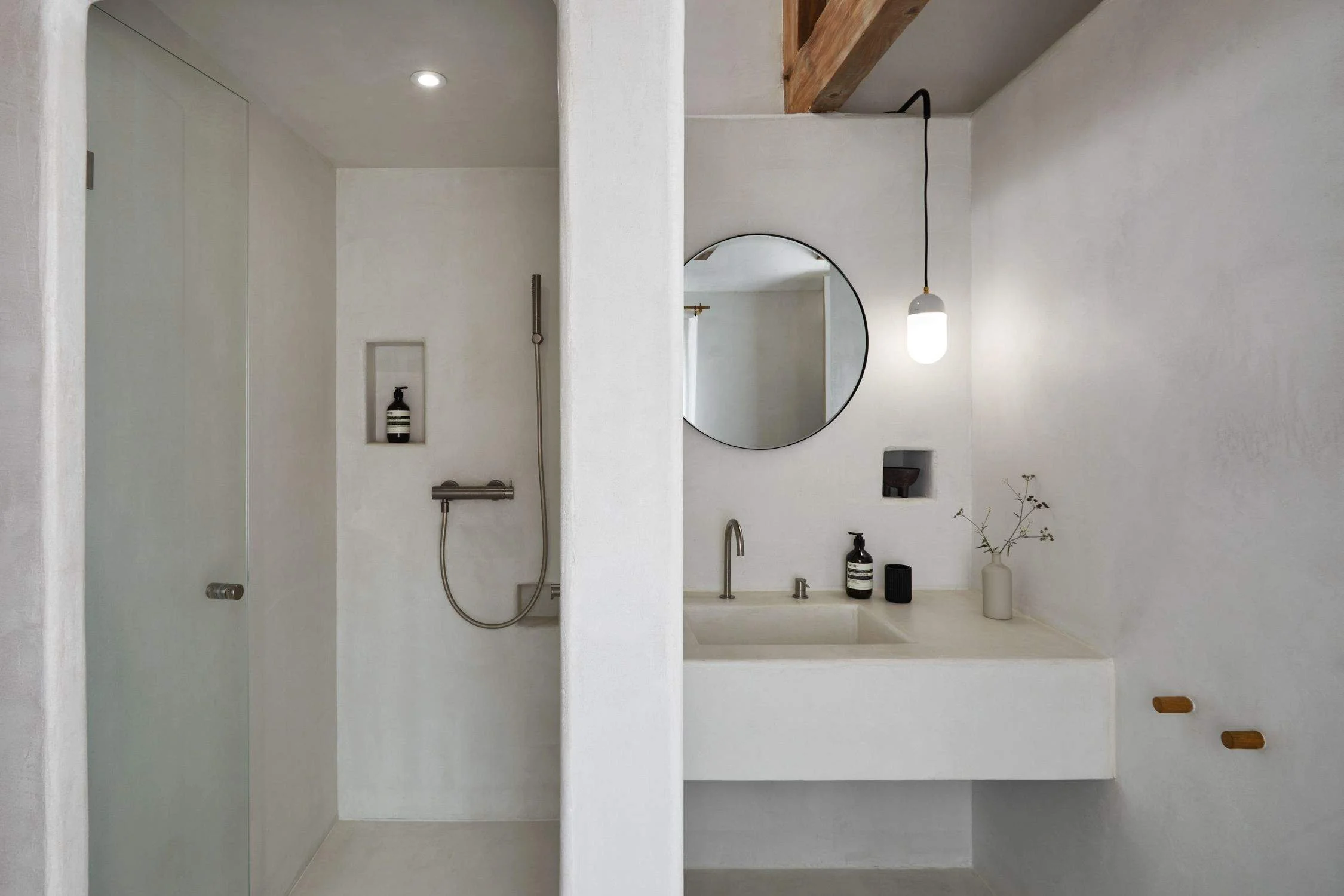 A minimalist bathroom with a walk-in shower on the left and a vanity on the right. The shower features a built-in niche with toiletries, a wall-mounted flexible shower head, and a glass door. The vanity has a round mirror, a modern faucet, a black soap dispenser, a black container, a small vase with dried flowers, and a pendant light hanging above.
