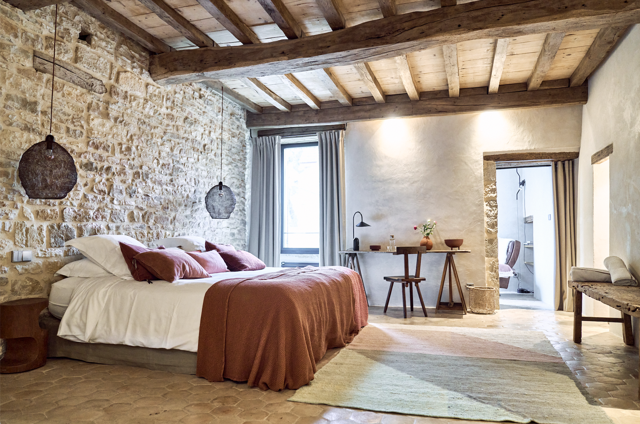A rustic bedroom with a stone wall, wooden ceiling beams, and a large bed with white and pink bedding. There are two hanging pendant lights, a window with curtains, a small wooden table with a chair, and an archway leading to a bathroom. The floor has a multicolored area rug.