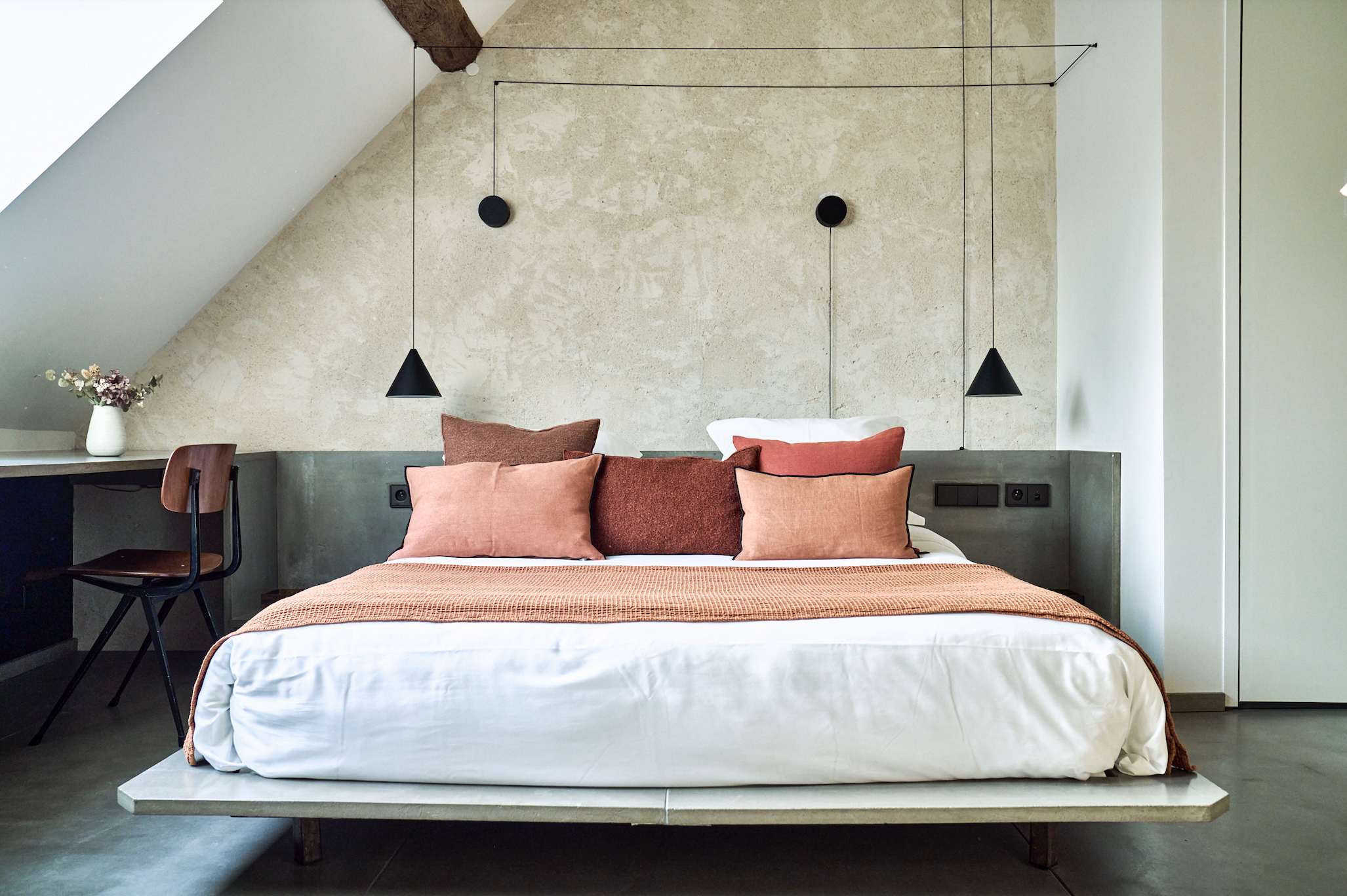 A modern bedroom with a bed covered in white and rust-colored pillows and a blanket, accented by black hanging pendant lights and a concrete wall backdrop.