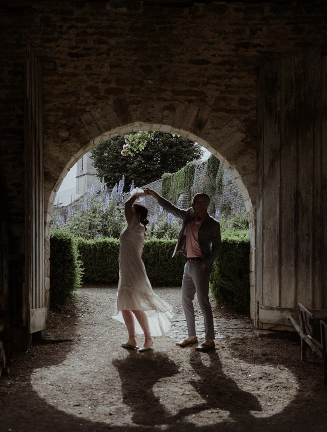 A couple dancing in a rustic stone archway, holding each other's hands and looking at each other, with greenery and a stone wall in the background.