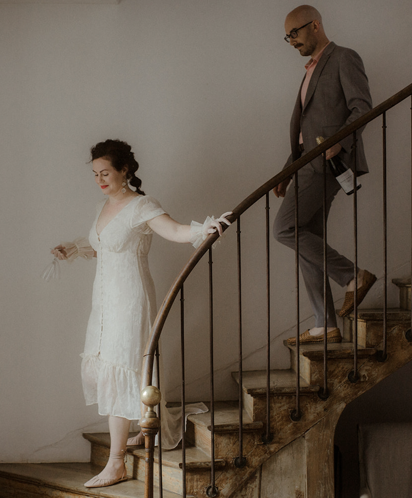 A woman in a white dress descending a wooden staircase, holding a flower, with a man in a gray suit and glasses carrying a bottle of wine following behind.