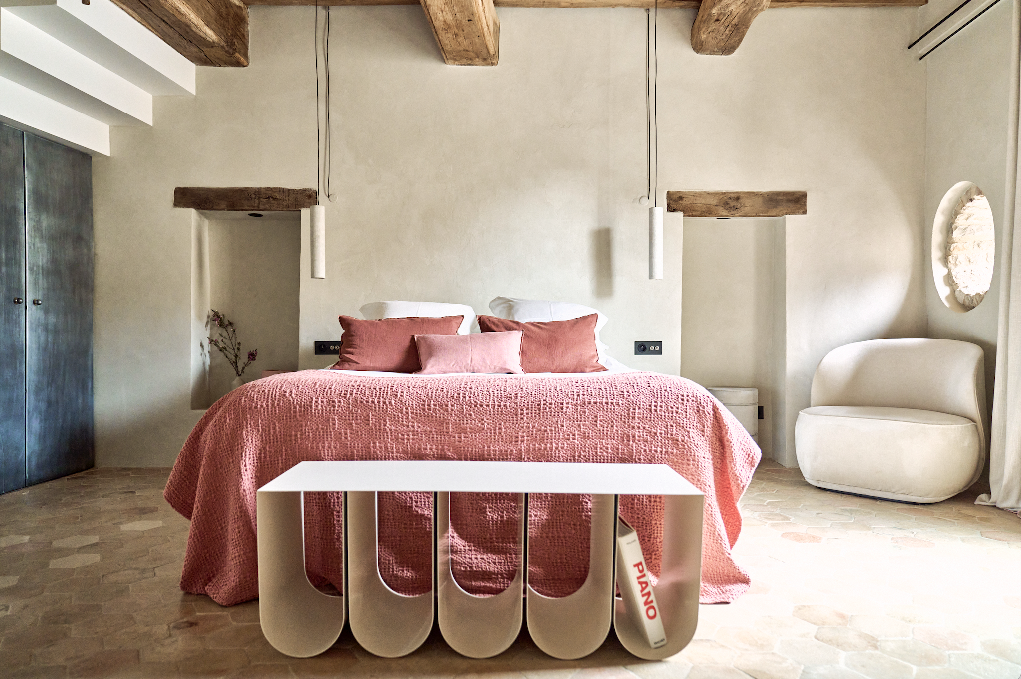Minimalist bedroom with a bed covered in a pink quilt and matching pillows, a white chair, a small table, and rustic wooden beams on the ceiling.