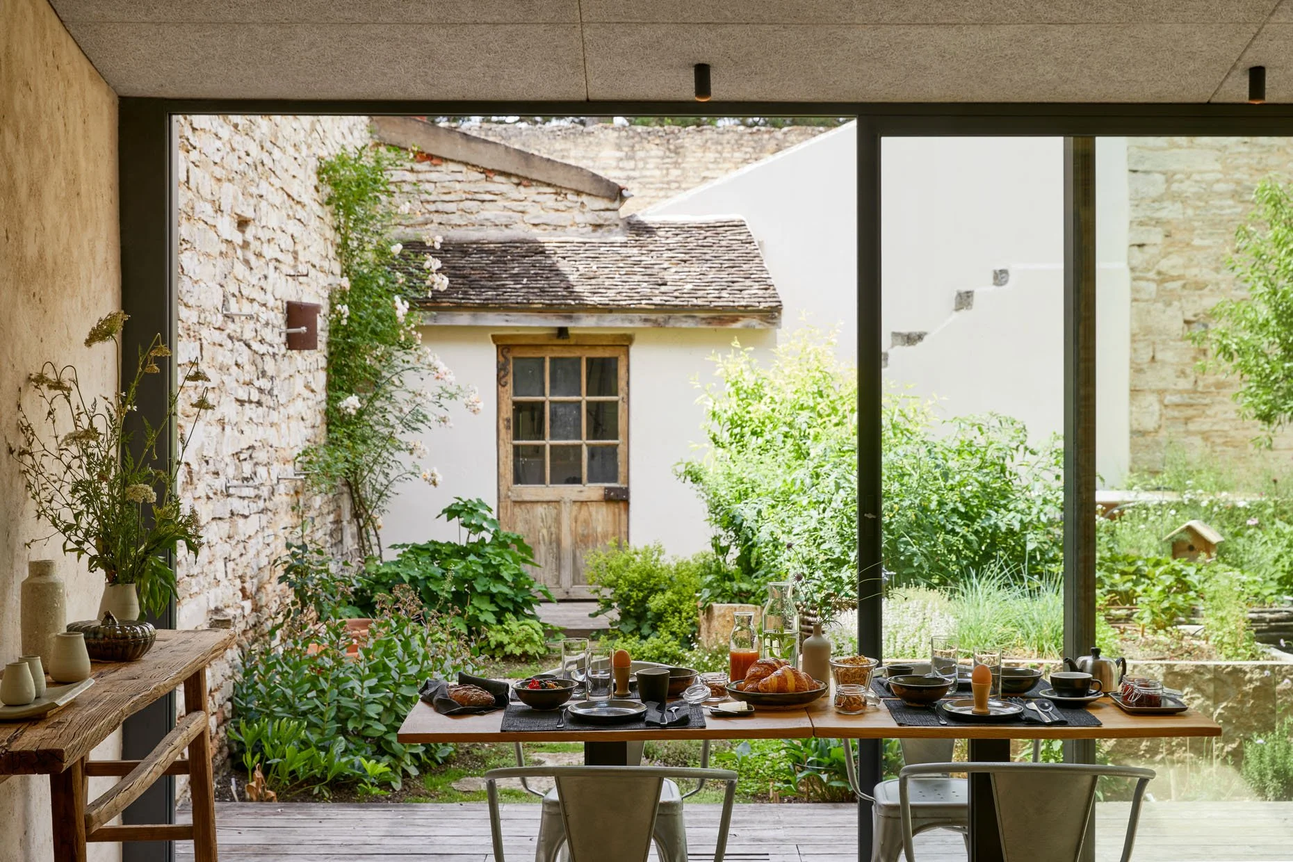 Breakfast table set with plates, bowls, glasses, and pitchers, overlooking a lush garden through a large glass window.