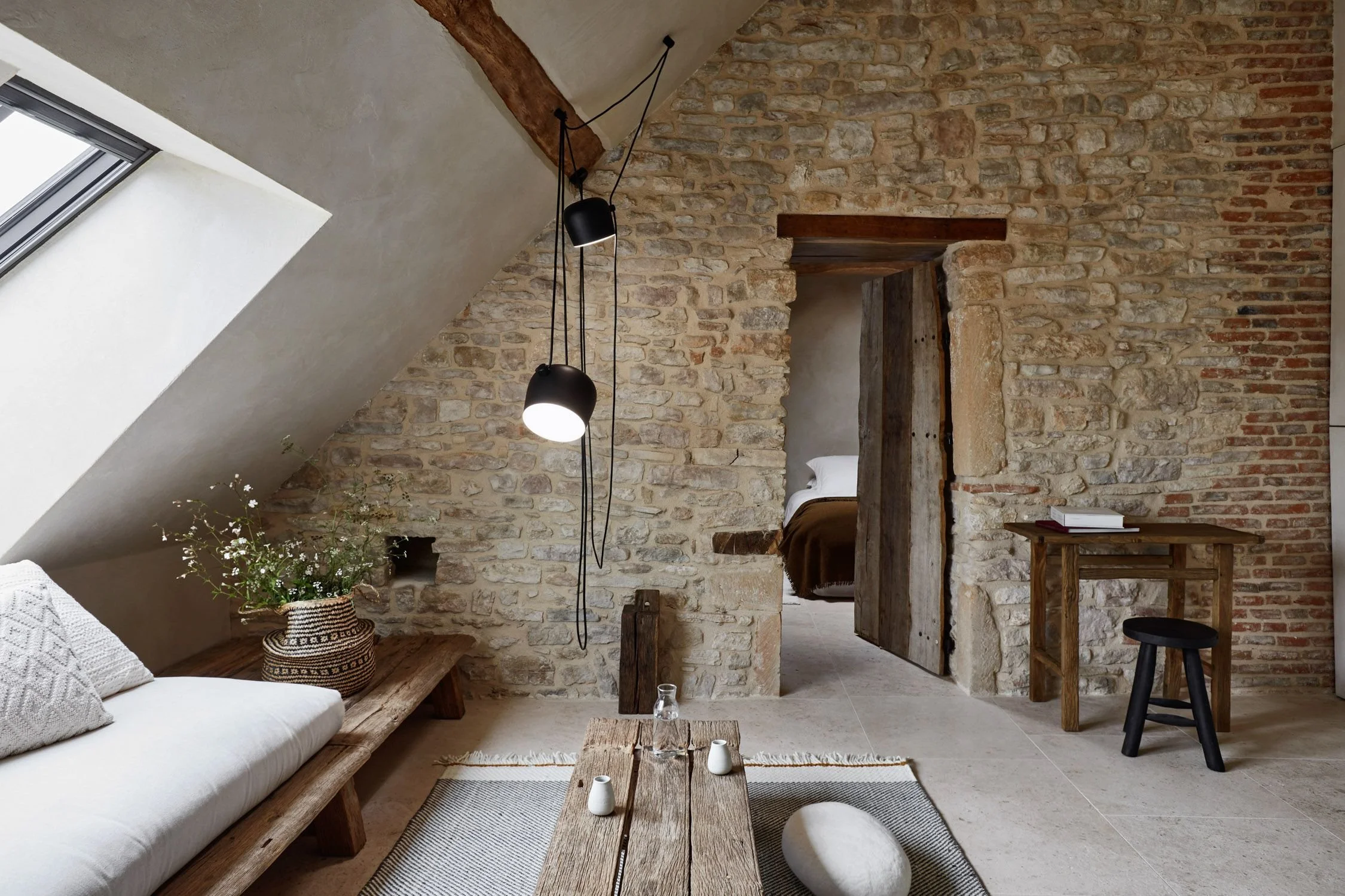 Interior of a rustic-style room with stone walls, a sloped ceiling, a skylight window, a white sofa with patterned pillows, a wooden coffee table with small vases, a potted plant, and a small wooden desk with a stack of books and a stool, leading to a bedroom with a bed and a barn door.