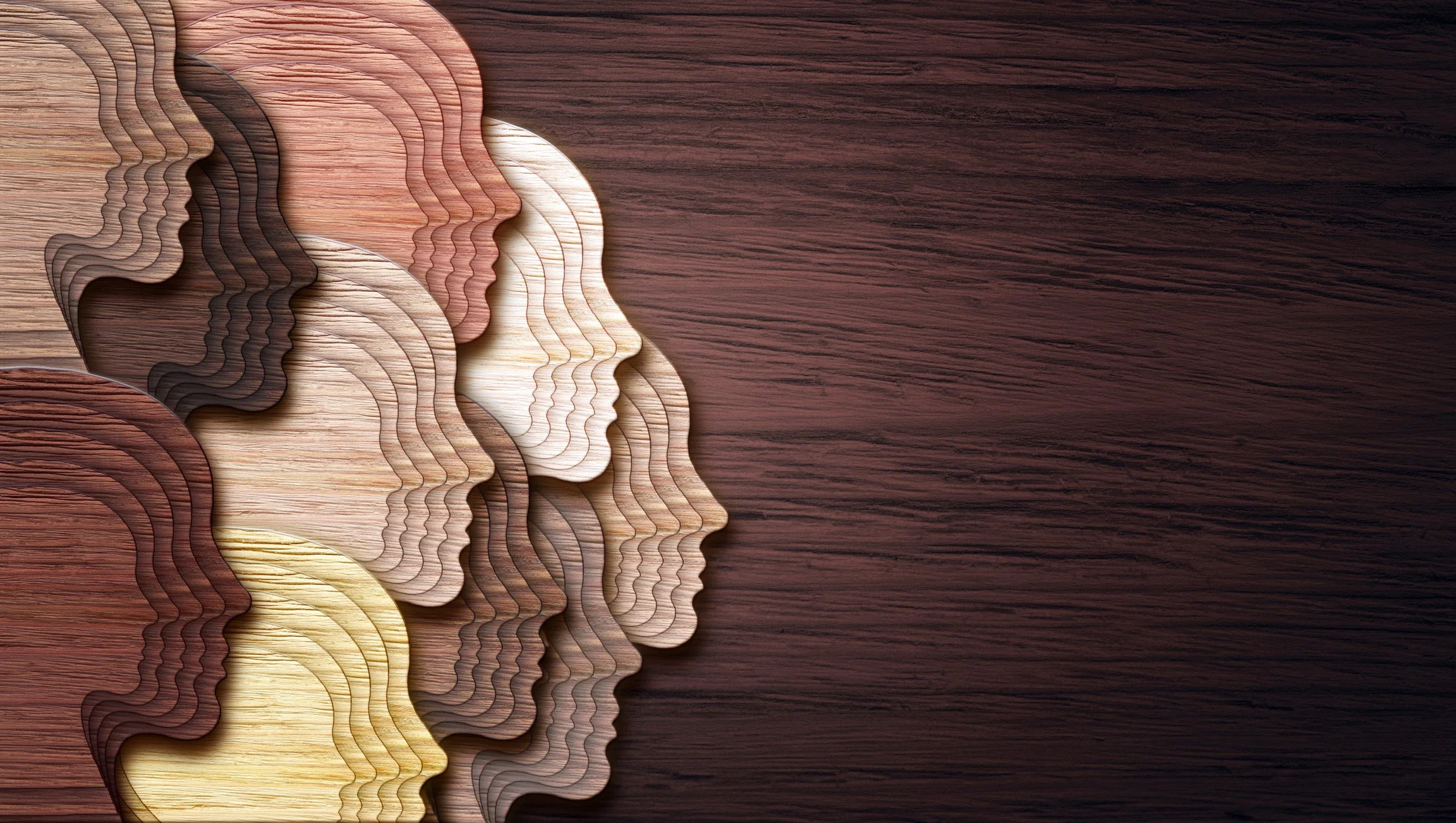 Multiple layered wooden silhouettes of human heads in various shades arranged on a dark wooden surface