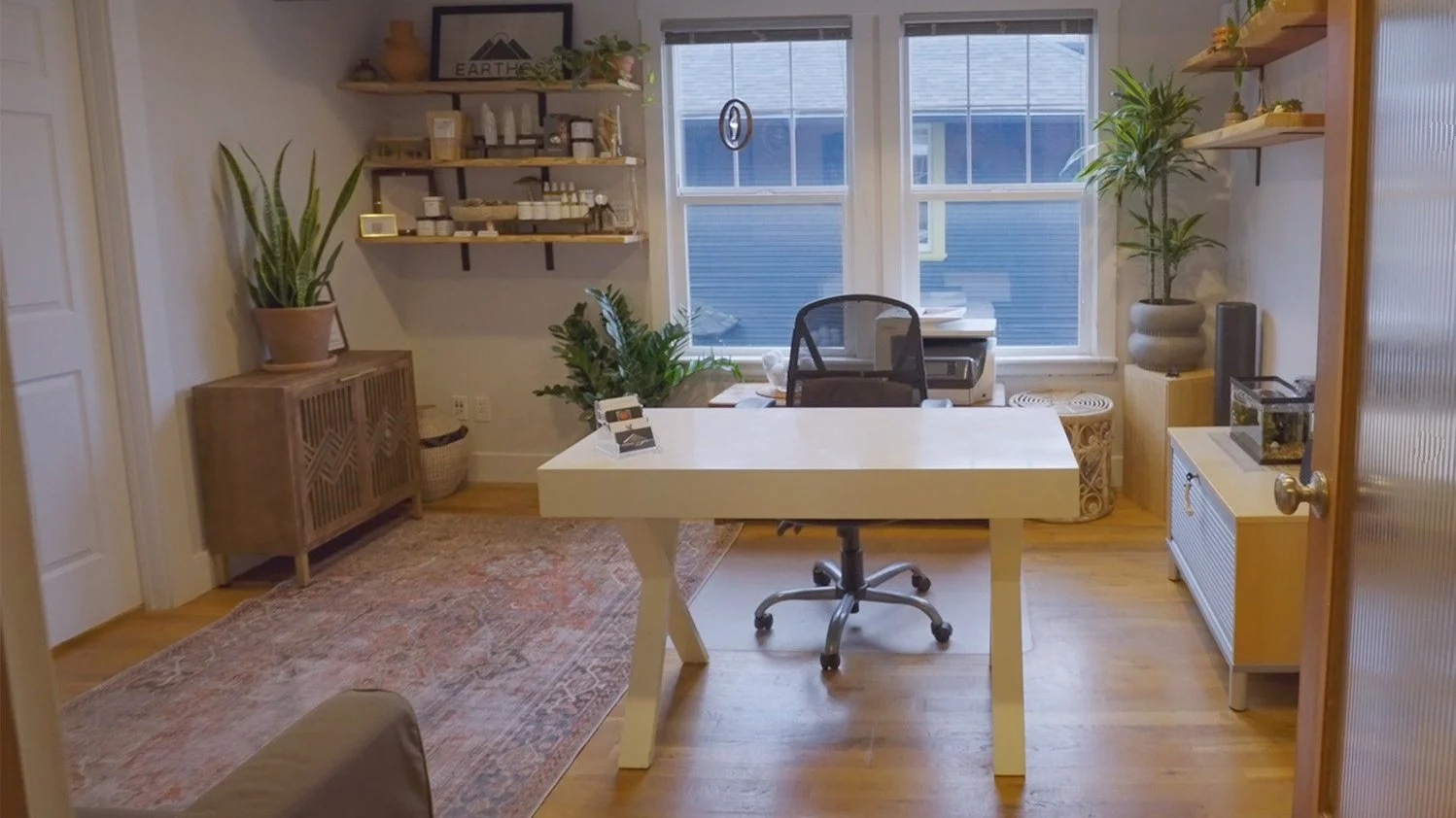 Bright home office with a white desk, black office chair, and large window, decorated with houseplants and shelves holding various items.