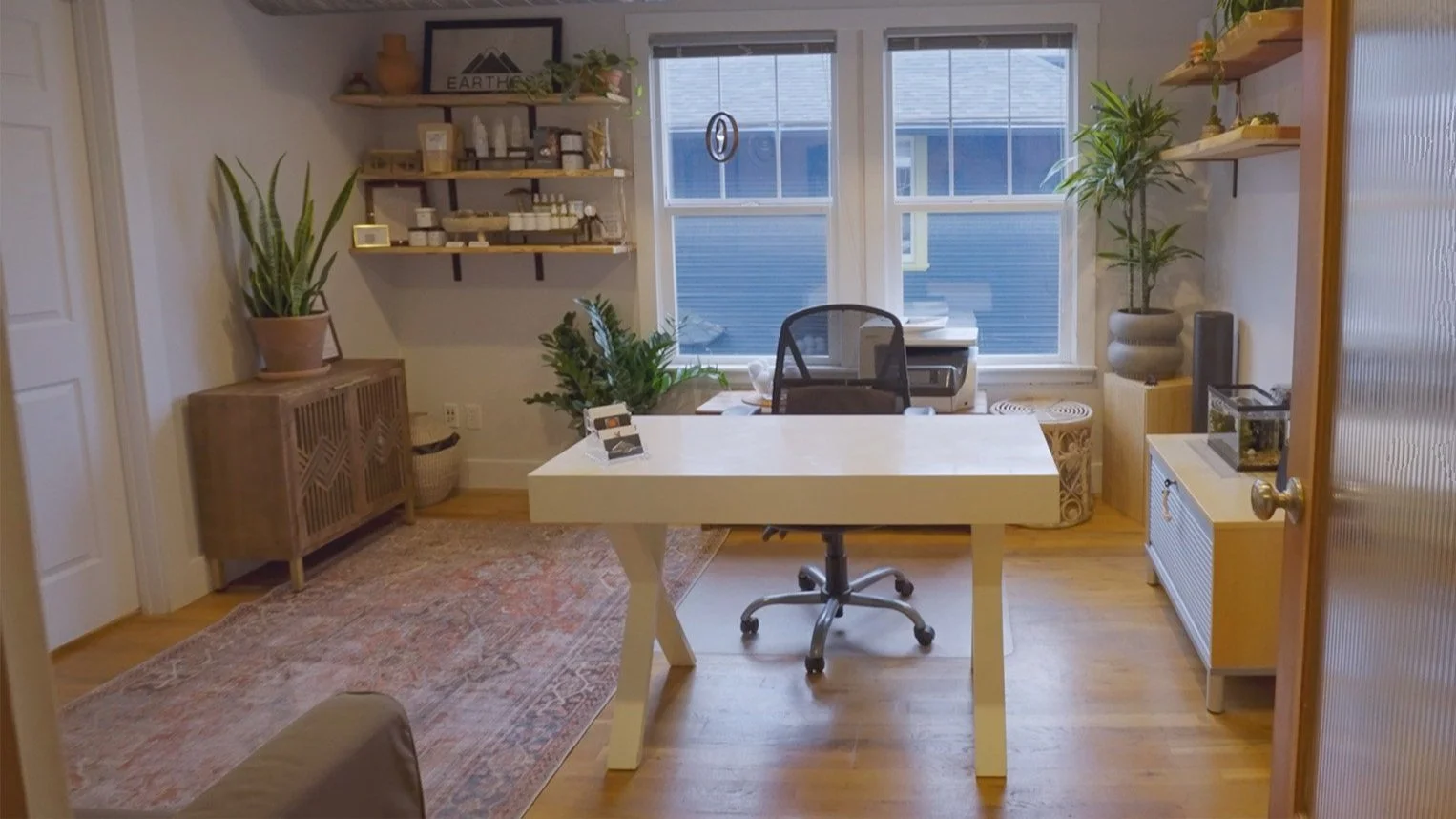 A bright home office with a white desk, black office chair, and large plant near the window. Shelves with decor and supplies, wooden sideboard, and a colorful rug on a wooden floor.