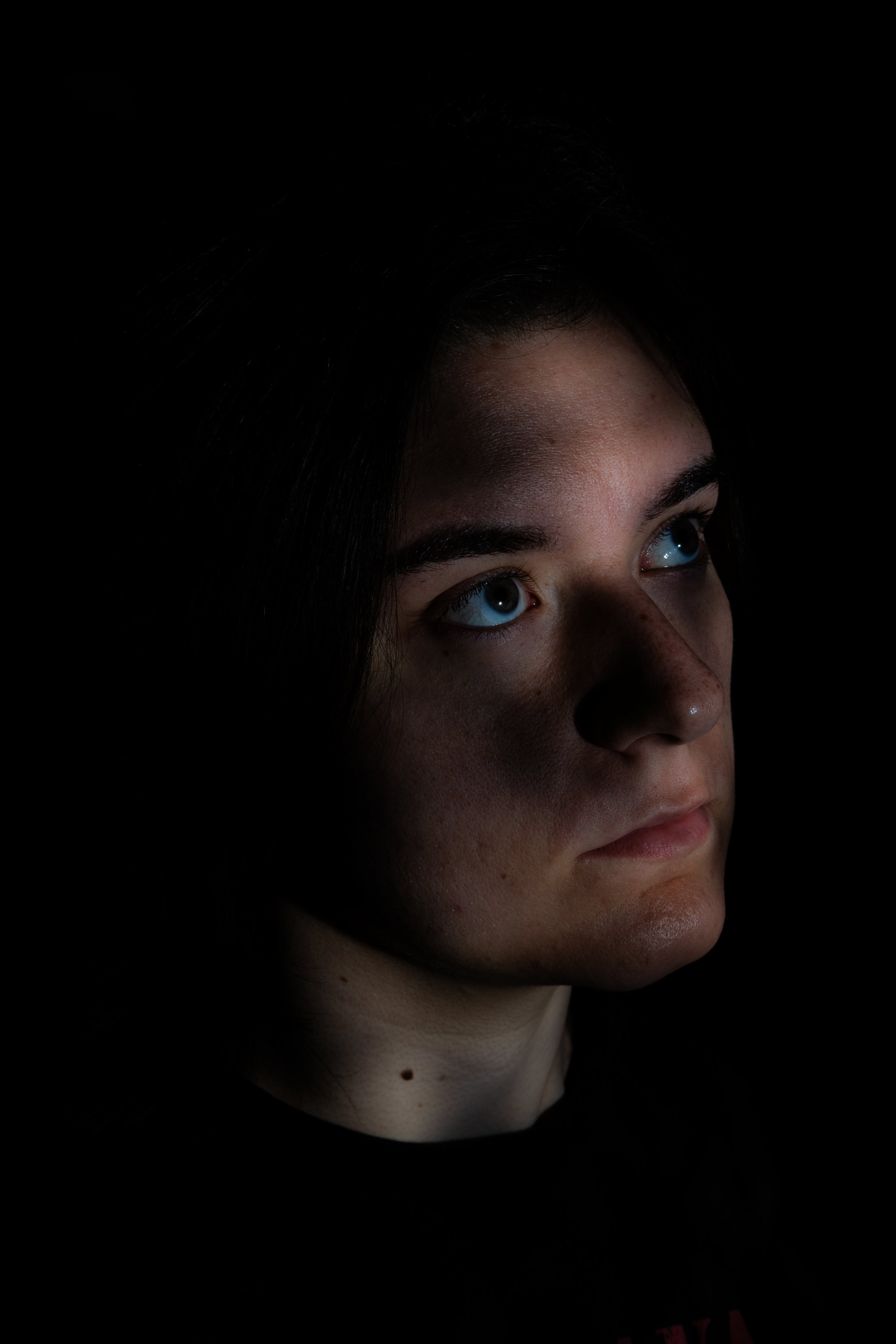 A young woman with green eyes and dark hair looks to the side in a dark setting with minimal lighting highlighting her face.