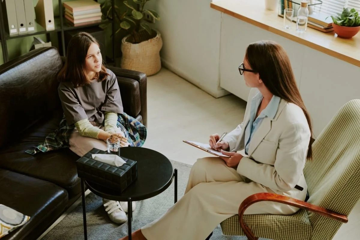 Young teenage girl sitting on a couch listening to her therapist taking notes during a youth therapy session.