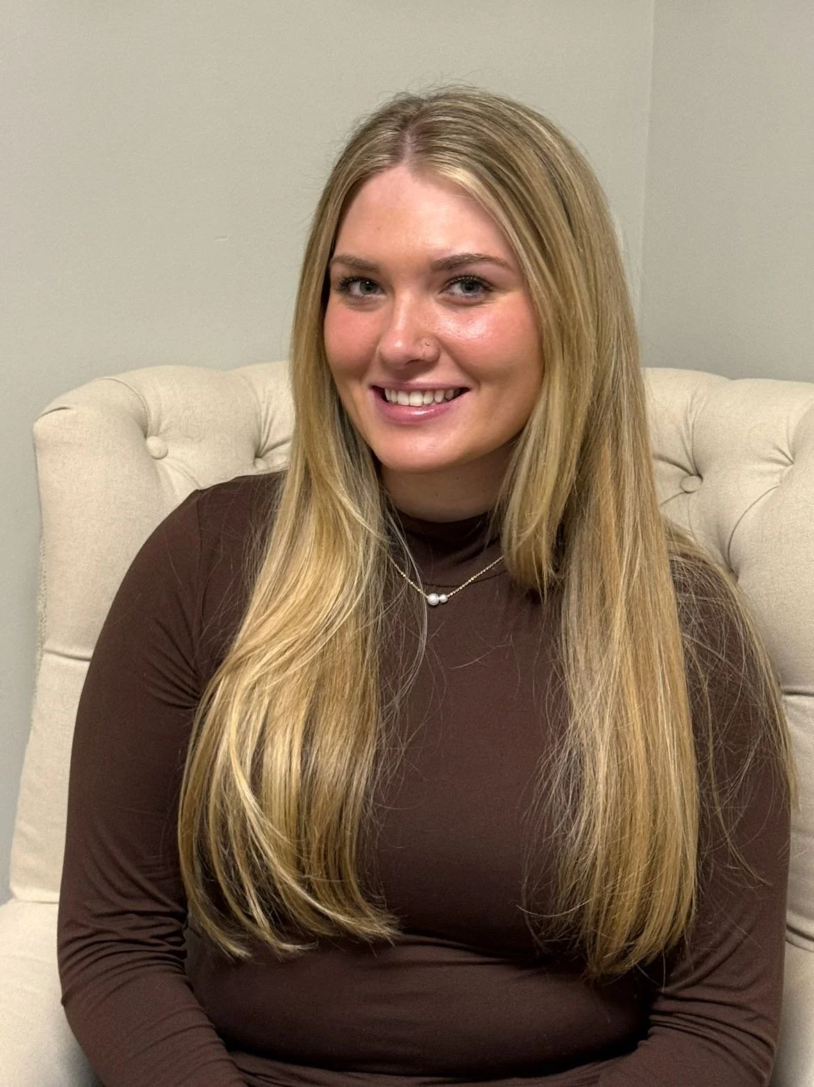 A young woman with long wavy blonde hair, light skin, wearing a black top, smiling against a plain gray background.