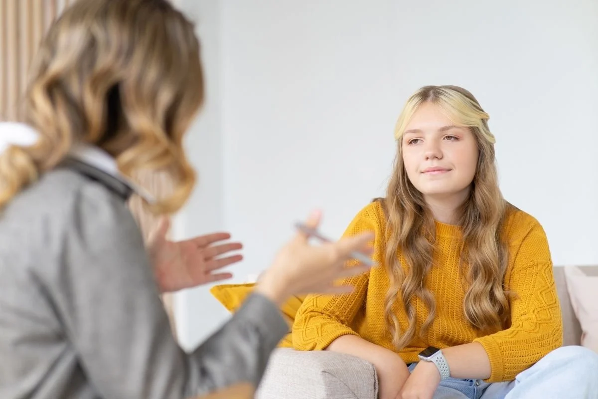 A teenage girl wearing a yellow sweater listening attentively to her therapist during a individual counseling session.