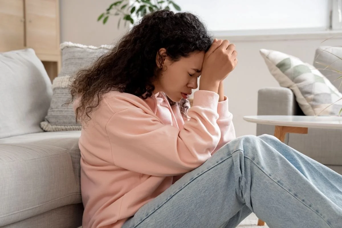 Young woman sitting on the floor leaning against a sofa holding her head during a panic attack or severe anxiety episode.