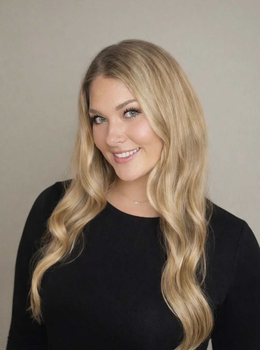 A young woman with long wavy blonde hair, light skin, wearing a black top, smiling against a plain gray background.