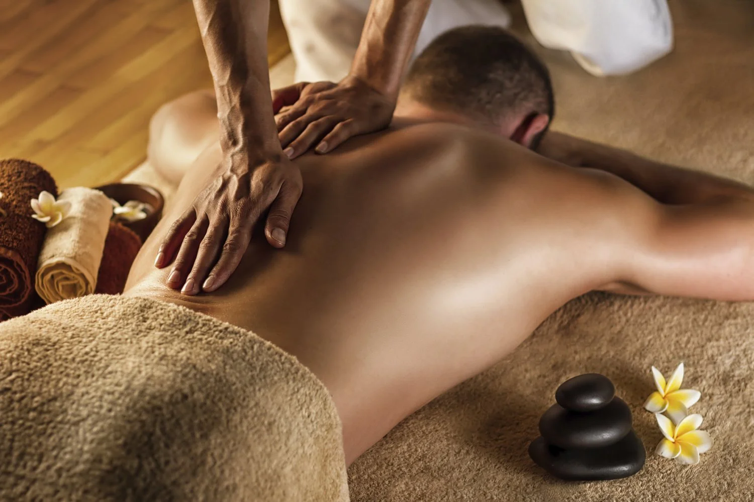 A person receiving a back massage in a spa setting with towels, flowers, and massage stones nearby.