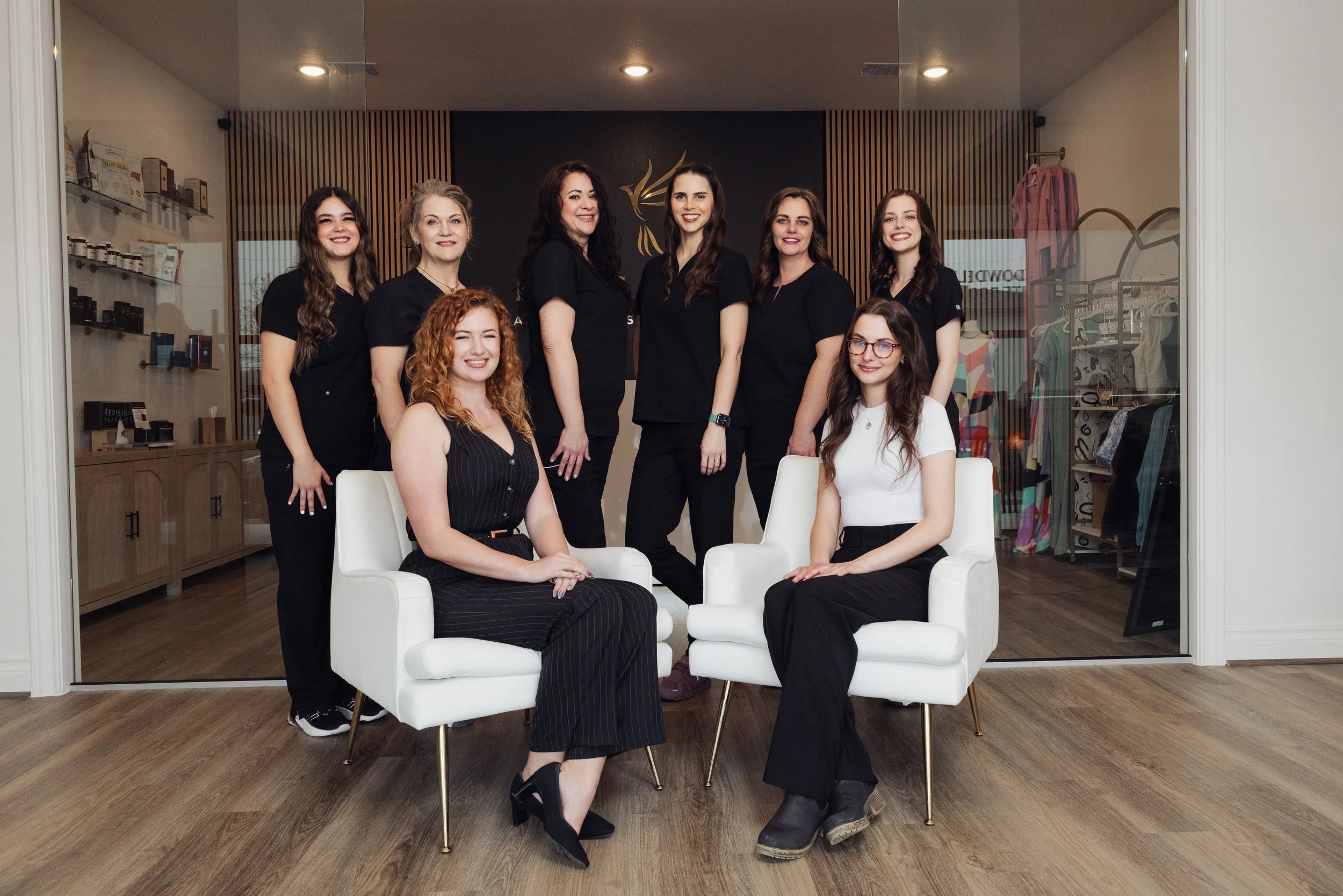 Group of nine women, some sitting on white chairs and others standing, inside a modern store or office space with wood flooring and display shelves in the background.