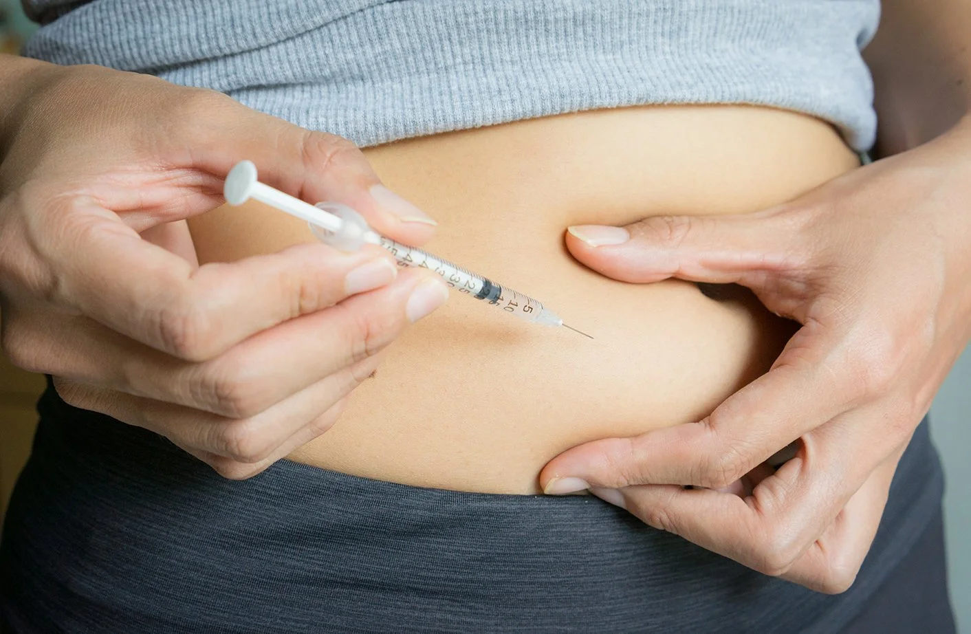 A person with a gray shirt pulls up their shirt to reveal their abdomen while another person is administering a weight loss shot using a syringe.