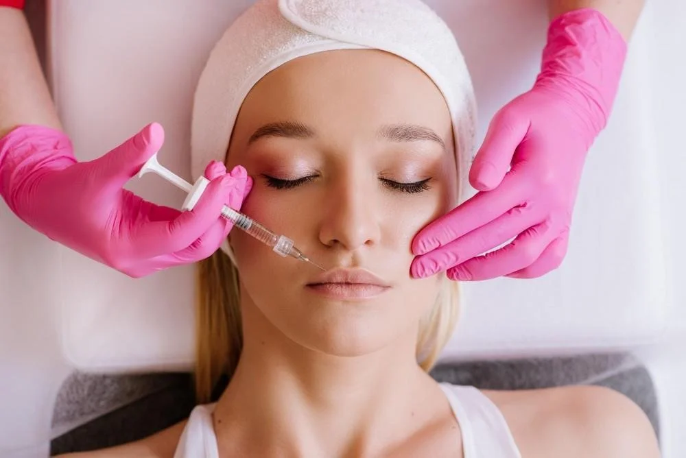 A woman receiving a facial cosmetic treatment, with a practitioner wearing pink gloves administering an injection near her eye.