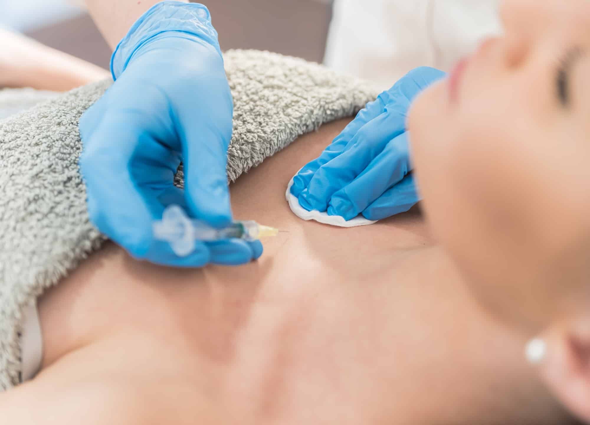 A healthcare professional in blue gloves administering an injection of Platelet Rich Plasma (PRP) into a woman's chest area for breast enhancement during a medical procedure.