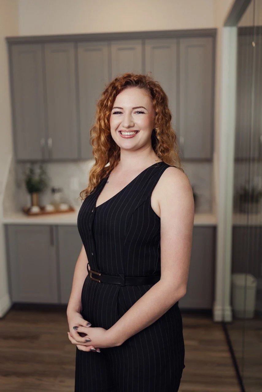 A smiling woman with curly red hair wearing a black pinstripe sleeveless dress standing in a modern kitchen.