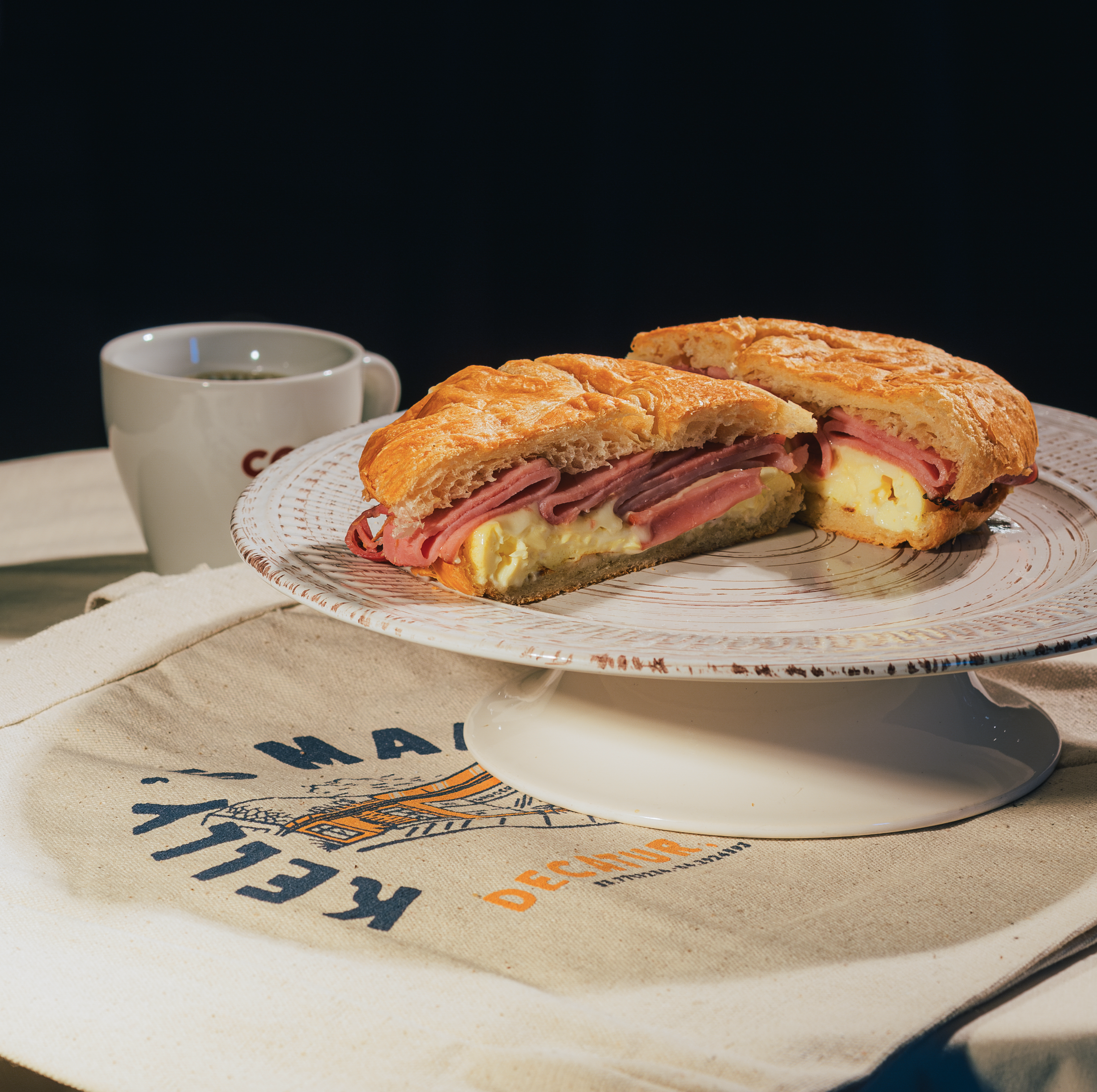A sandwich filled with ham, cheese, and eggs, cut in half and served on a decorative plate, with a cup of coffee in the background.