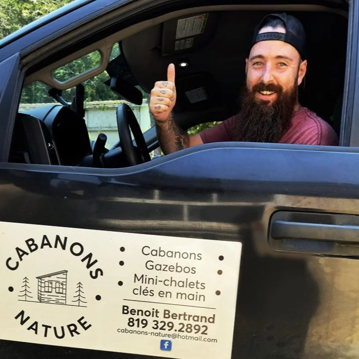 Cabanons nature, cabanon Outaouais. Homme souriant avec barbe et chapeau de baseball, donnant un pouce en l'air depuis le véhicule, fenêtre ouverte.