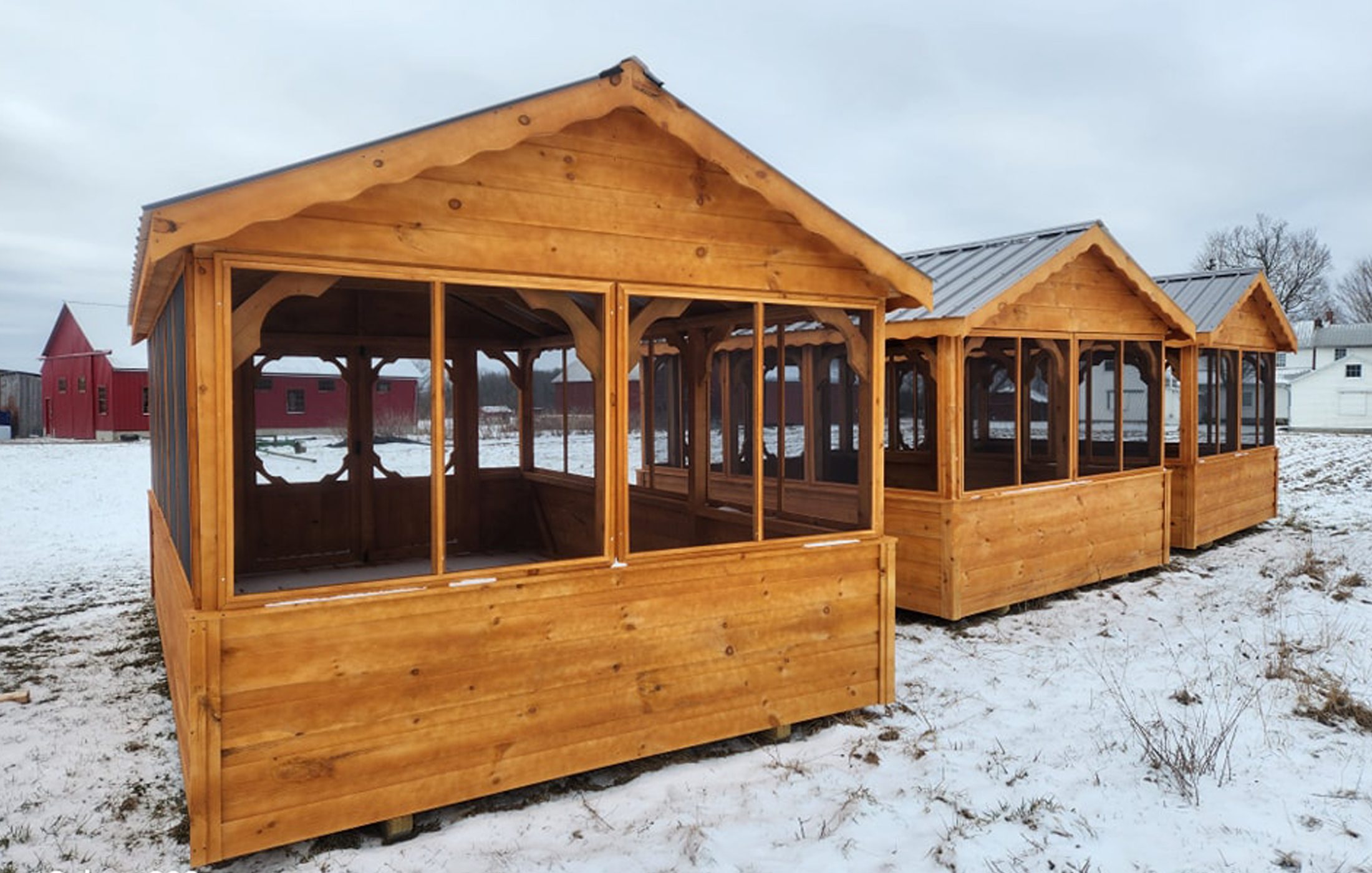 Cabanons nature, cabanon Outaouais. Gazebo en bois traité, en extérieur sous un ciel nuageux, avec neige au sol, maisons blanches et rouges en arrière-plan.