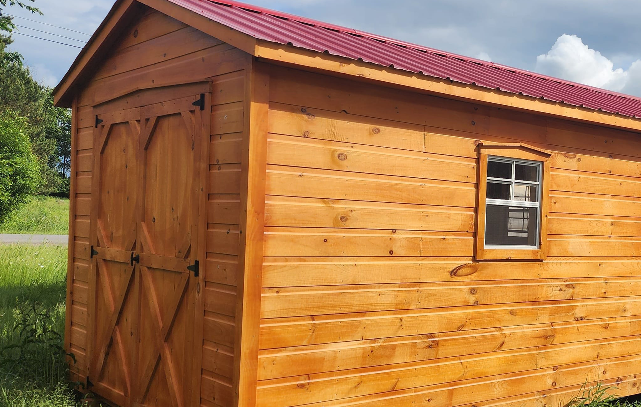 Cabanons nature, cabanon Outaouais. Un cabanon en bois avec des murs en planches traitées, une porte double en bois avec des ferrures noires, une fenêtre à petits carreaux, et un toit en métal rouge, Cabanon. Backyard Shed. Remise. Cabanons.