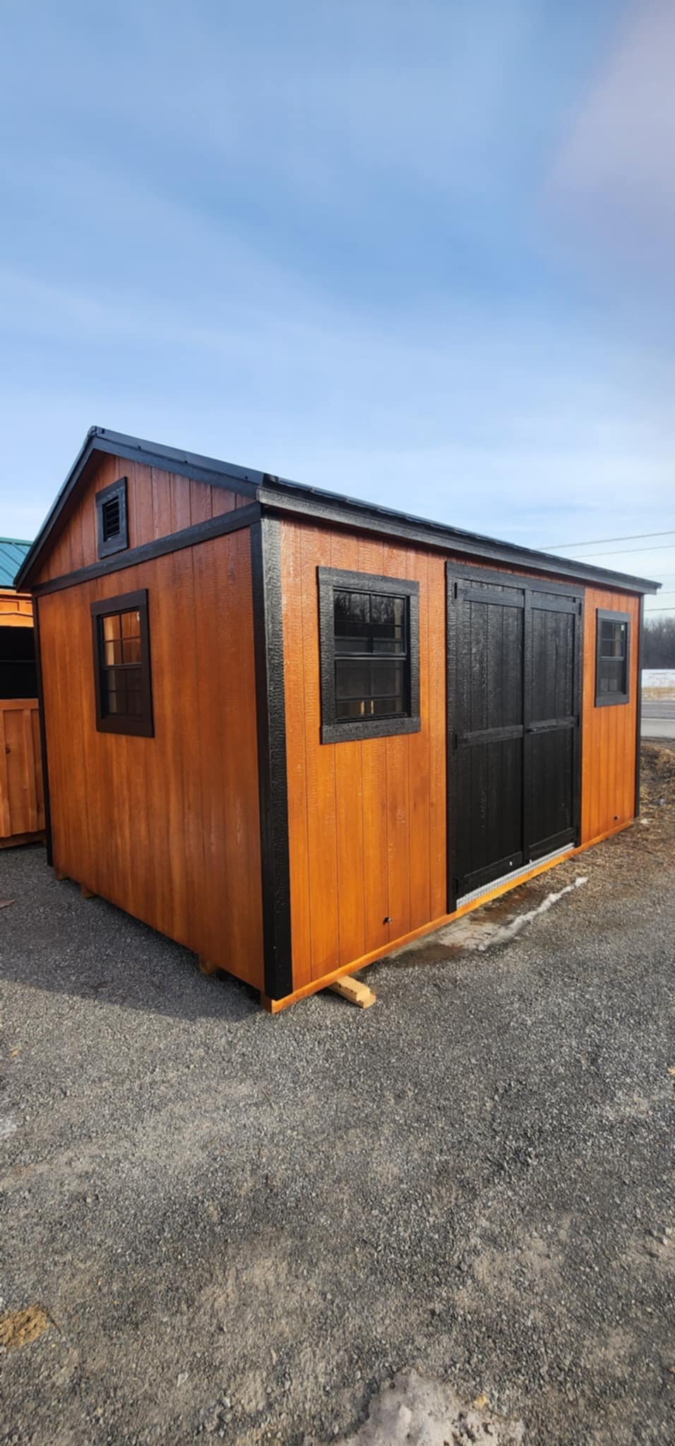 Cabanon Outaouais, Cabanons Outouais. Petite cabane en bois avec double porte noire et fenêtres à encadrement noir, posée sur un sol en gravier.