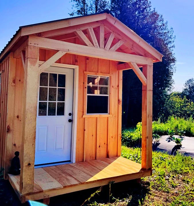 Cabanon Outaouais, Cabanons Outouais. Petite cabane en bois avec une porte blanche à fenêtre, sur une terrasse en bois, dans un environnement de jardin ensoleillé. Cabanon. Backyard Shed. Remise. Cabanons.