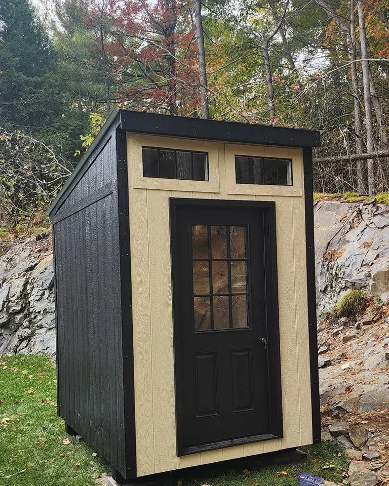 Cabanons nature, cabanon Outaouais. Une petite cabane en bois avec une porte noire et deux petites fenêtres en haut, située dans un environnement naturel avec des arbres et des rochers en arrière-plan. Cabanon. Backyard Shed. Remise. Cabanons.
