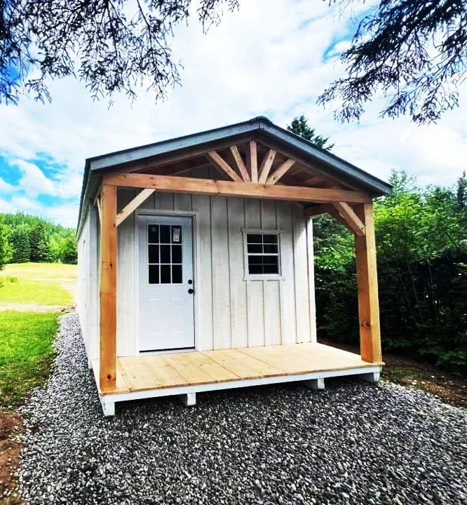 Cabanon Outaouais, Cabanons Outouais. Petite cabane en bois avec un porche, située sur un sol en gravier, avec un ciel bleu et des arbres en arrière-plan. Cabanon. Backyard Shed. Remise. Cabanons.